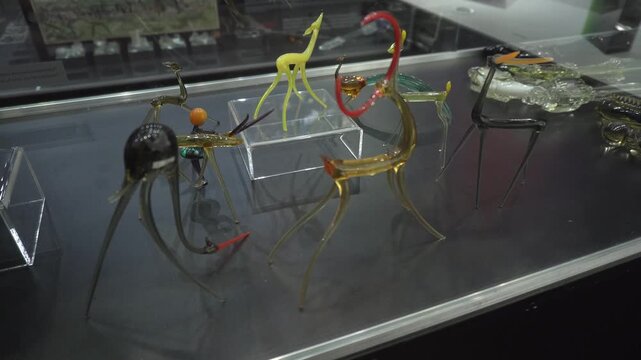 Assortment of vibrant transparent glass animal sculptures featuring elongated forms and intricate details, presented under protective glass in a dark setting, highlighting artistry of each unique piec