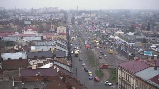 Elevated perspective of a vibrant city thoroughfare featuring continuous flow of vehicles, including buses and private cars, with tram lines visible. Urban landscape extends to horizon under gray, clo