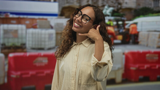Woman smiling with hand to ear phone gesture at outdoor building construction site wearing glasses and casual shirt; friendly invitation.