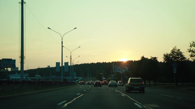 Viewpoint from inside car of urban road at dusk, sun setting on horizon, creating beautiful golden light across sky and trees, vehicles in motion, journey on busy street