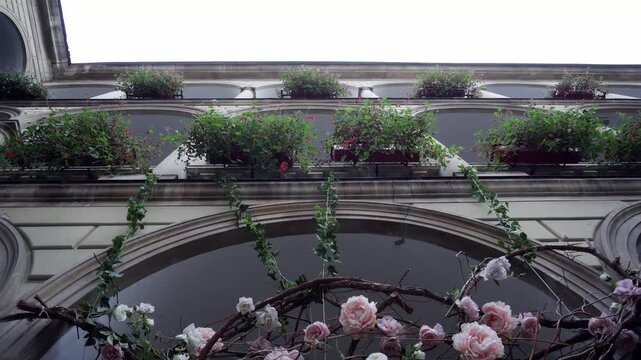 Building facade, multiple arched levels. Lower arch with pink and white roses, branches, vines. Upper balconies, flower boxes, green plants, red flowers. Ornate architecture, viewed from below.