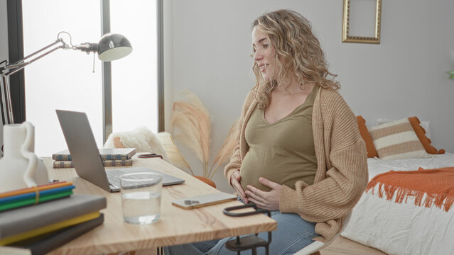 Pregnant woman holding belly at laptop in bedroom, smiling and cradling bump beside bed and desk while seated; contentment.