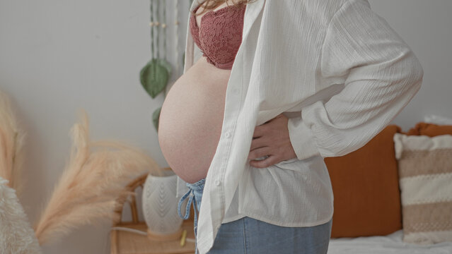 Woman showing pregnant belly, hand on lower back supporting abdomen in bedroom; expectant calm maternal intimacy.
