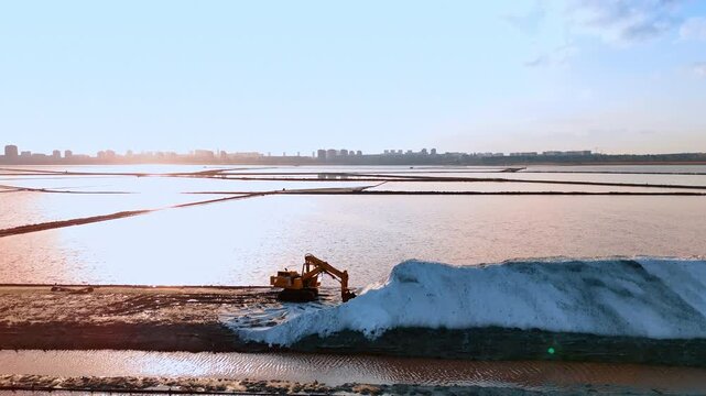 Industrial excavator working at salt evaporation pond during sunset. Heavy machinery moving white salt pile at a coastal salt farm with sun reflection on water.