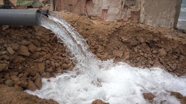 Close-up of a heavy water stream discharging from a plastic pipe into a rocky dirt basin, with makeshift housing in the background. Ideal B-roll for poor infrastructure and wastewater runoff.