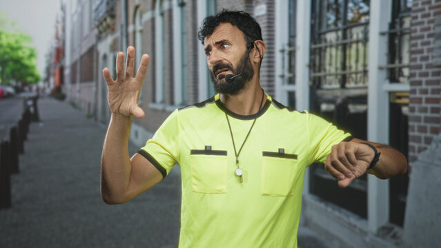 Referee man with whistle and raised hand checks wristwatch on cobbled street by building facade, headset visible; time pressure concern.