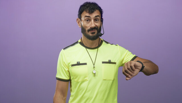 Man referee checking his watch and pointing finger, whistle on lanyard and headset mic visible, yellow referee jersey against purple studio wall; urgency.