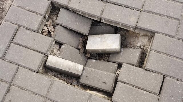 Close-up aerial view of significant damage to walkway surface, revealing multiple gray paving blocks dislodged and collapsed into a deep cavity, exposing underlying soil and debris.