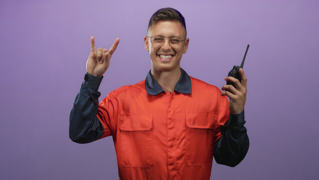Young mechanic man wearing orange coverall and glasses, smiling and holding a walkie talkie while flashing rock hand sign in studio; playful joy.