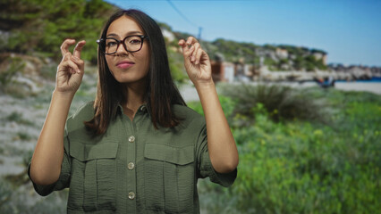 Naklejka premium Young hispanic woman with glasses and brown hair making air quotes gesture in building near a green park and coastal dunes; playful skepticism.