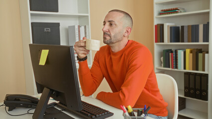 Man in orange sweater holds mug with both hands at computer desk among bookshelves in building, gazing at screen  quiet contemplation. © Krakenimages.com