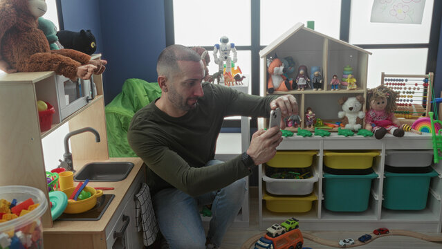Man holding smartphone taking a photo while kneeling by toy shelves and a play kitchen in a building kindergarten room with dolls and bins; warmth.