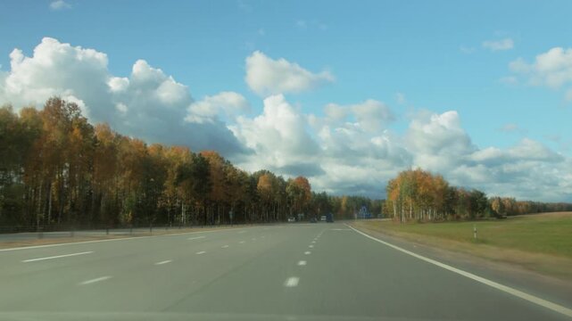 Driving view on wide country highway on bright autumn day. Vibrant fall foliage lines roadside. Speed limit and warning signs visible. Few cars on asphalt road under blue sky.