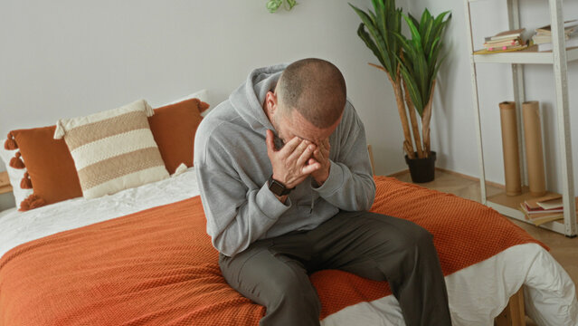 Man on bed sitting covers face with hand in a building bedroom, gray hoodie and watch on wrist visible; sadness fatigue.