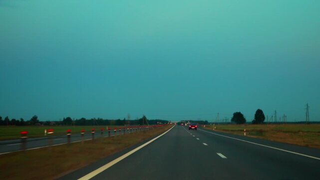 View of a multi-lane highway during evening twilight. Road markings guide path forward with car tail lights visible in distance. Surrounding landscape consists of fields, trees, and power lines under 
