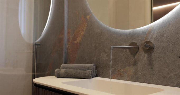 Luxury bathroom panorama with stone vanity wall and rounded lit mirror, smooth camera pan reveals wall mounted faucet with flowing water gray towels ribbed wood cabinet diffuser soap dispenser