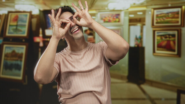 Man smiling with hands forming circular frames over his eyes, wearing a pink t shirt and standing before framed paintings in an art gallery building; playful.
