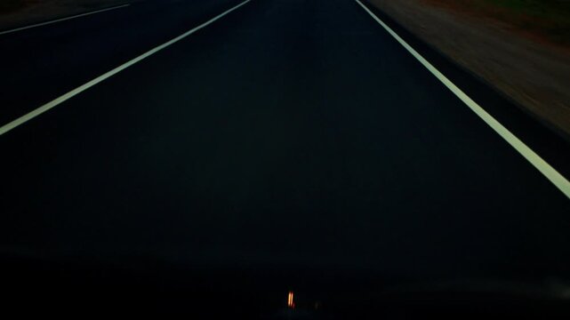 Dark asphalt road stretches forward into distance, illuminated by faint light and marked by white lines. Scene depicts long journey and solitude of night travel by vehicle