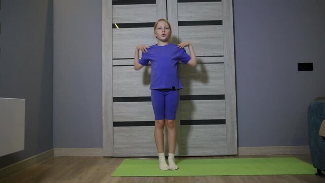 Full length view of young blonde girl performing shoulder circles on green mat in modern home environment. Child focused on physical activity, promoting healthy lifestyle for kids wellbeing