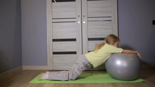 Small girl engaged in active physical training, kneeling on a yoga mat and pushing a large fitball, promoting healthy lifestyle and proper body development