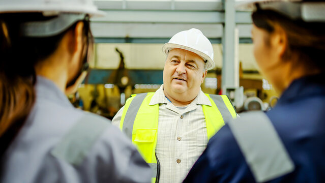 Senior male supervisor in hard hat and safety vest talking to female engineers in a modern factory. Industrial teamwork, heavy manufacturing site leadership, and professional worker coordination.