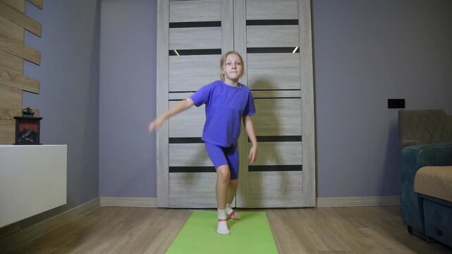 Young girl in blue sportswear performs lateral steps with resistance band. Exercise targets leg muscles. Child trains on green yoga mat at home.