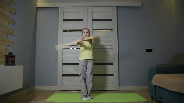 Young blond boy performs martial arts weapon training indoors. Child rotates long wooden stick horizontally with extended arms. Focus and concentration shown on green mat.