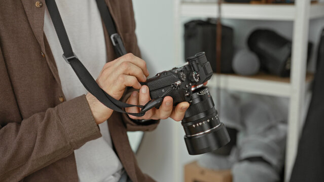 Young man adjusts camera dials while holding lens and strap in a photography studio with shelves of gear and softboxes visible; concentration craft.