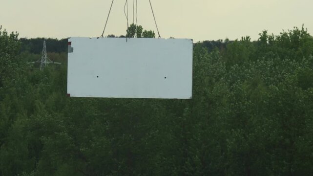 White rectangular panel suspended by chains from crane over green forest canopy. Industrial lifting operation transport building material. Power line tower visible.