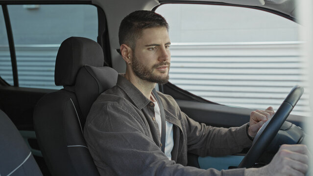 Man driving with hands on steering wheel inside car beside corrugated metal building facade, gazing forward in a steady pose; calm contemplation.