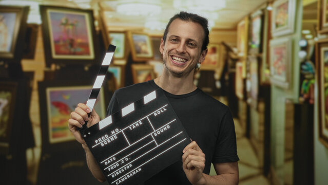 Man with long hair holding a clapperboard in an art gallery building, tilting head and smiling while raising the clapper; playful creativity.