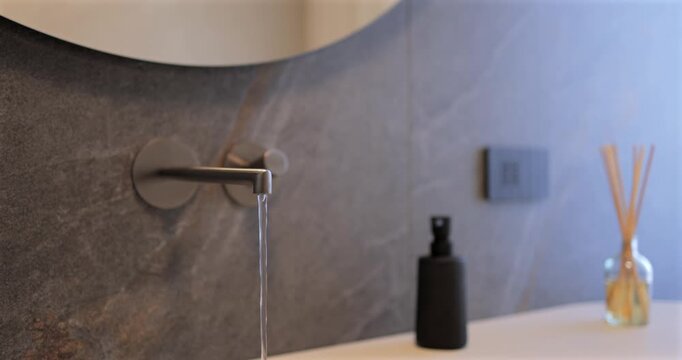 Natural stone washroom detail with close focus on sleek built in spout and flowing stream, still footage holds on quiet composition as daylight from window wraps sink area beside round mirror