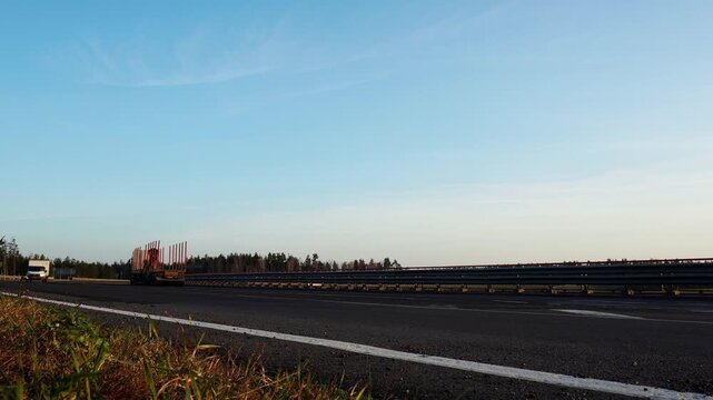 Long exposure view of moving logging truck carrying timber on multi-lane highway, clear blue sky above, forest line background, transport infrastructure and supply chain activity