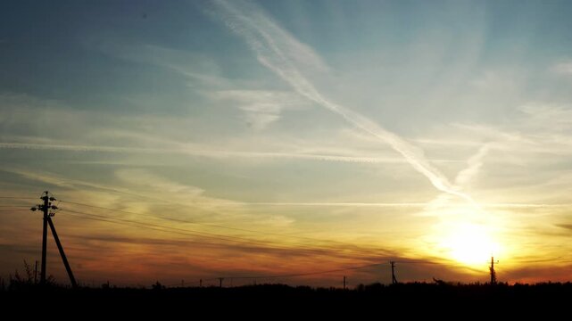 Large commercial truck in silhouette driving on highway at evening. Sunset sky with striking orange and yellow hues. Prominent airplane contrails across blue sky. Utility poles on horizon.