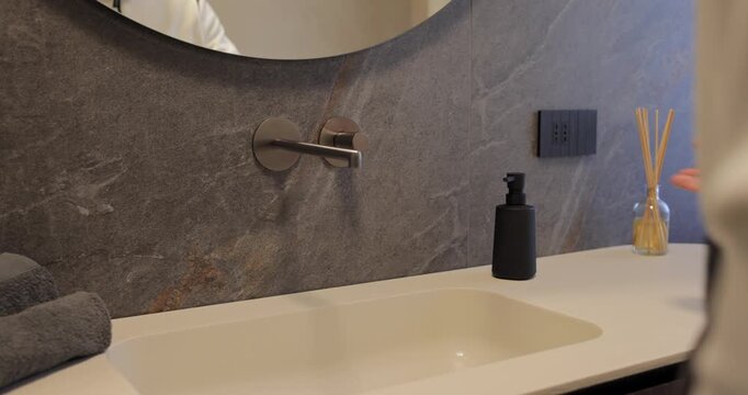 Premium washroom detail with touchless mirror lighting above a pale basin, still footage captures a hand passing under the circular edge as soft illumination spreads across textured stone and decor