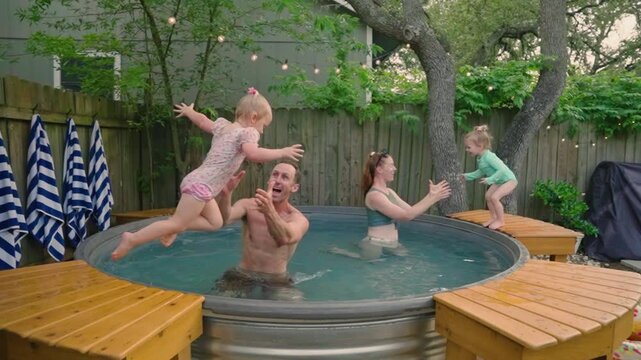 Family splashing in a backyard stocktank pool, dad lifting one twin and mom holding the other while all four laugh and play. Perfect for family lifestyle ads, summer fun, and pool day content.