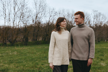 Fototapeta premium Couple outdoors strolls through a quiet park in knit sweaters, sharing warm smiles and gentle contact, enjoying a calm lifestyle amid bare trees and open fields, romantic connection