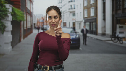 Fototapeta premium Woman pointing finger at camera on a quiet urban street, wearing maroon long sleeve sweater, jeans and belt, earrings visible; confidence direct address.