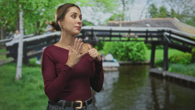 Woman in burgundy off shoulder shirt touching chest and clasping hands near collarbone, looking upward in studio with canal backdrop; calm reflection.
