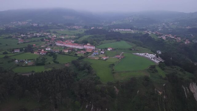 Aerial view of the village of Cudillero surrounded by lush green hills and misty forests under an overcast sky in Cudillero, Asturias, Spain.
