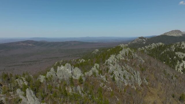 Drone footage of rocky mountain ridge and vast taiga forest under blue sky in Taganay National Park near Zlatoust, Chelyabinsk Oblast, Russia