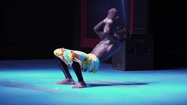 Powerful black African man with bare torso executes dynamic acrobatic dance performance on blue stage. Body contorts mid-air, showcasing incredible strength, flexibility, and artistic expression under