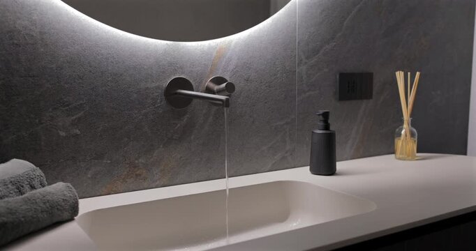 Running water from sleek wall faucet beneath glowing mirror in premium washroom, still interior footage focuses on steady stream over light basin against textured stone with elegant decor