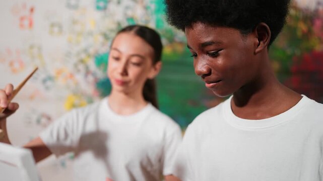 Diverse happy children paint canvas together with colorful stained wall. Smart multicultural student drawing together in art lesson while wearing white shirt. Creative activity concept. Edification