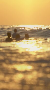 Group of people swimming in ocean at sunset or sunrise, warm golden light reflected on water surface, soft focus foreground with sparkling bokeh, relaxed summer vacation activity. Vertical shot. Summe