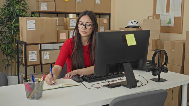 Woman types at computer amid stacked cardboard parcels and reaches for a pen in building; productivity calm.