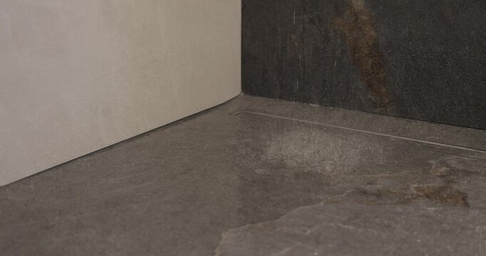 Slow motion shower droplets striking dark stone floor in modern wet room, close video captures splashes and ripples around linear drain as water falls from above in premium minimalist bathroom setting