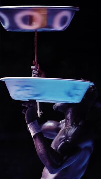 African male performer executes balancing act. Bowl on head, second bowl on stick in mouth, third bowl atop stick. Dark stage, dramatic lighting highlights muscular physique, concentration, skill. Ver