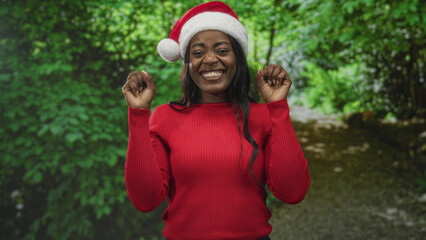 Naklejka premium Woman in santa hat and red sweater makes claw hands gesture while smiling broadly in forest setting, facing camera; holiday celebration joy.