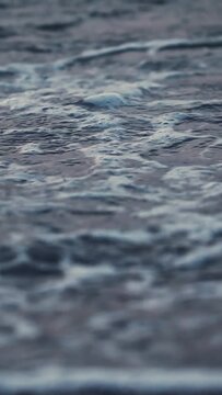 Close-up detail of ocean water gently breaking on shore line, creating foamy white wave crests receding across dark textured pebbles. Calm rhythmic movement of nature. Vertical shot